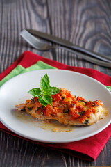 Grilled chicken breast with tomato and mozzarella cheese on it. On plate on wooden table with red and green napkins with fork and knife, vertical