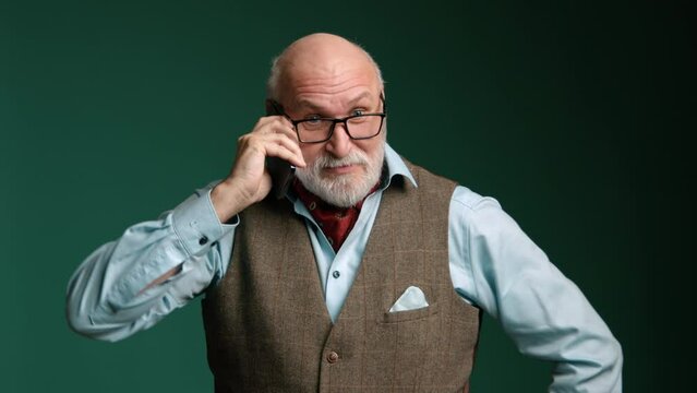 Dressed in a vest and light blue shirt, a man expresses shock and frustration during a contentious phone call on a deep green background. Camera 8K RAW. High quality 4k footage