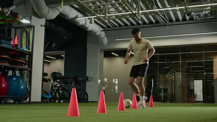 Caucasian sport man footballer soccer player running with ball feinting dribbling between cones playing football run fast motion feint on field in gym male guy sportsman professional training workout