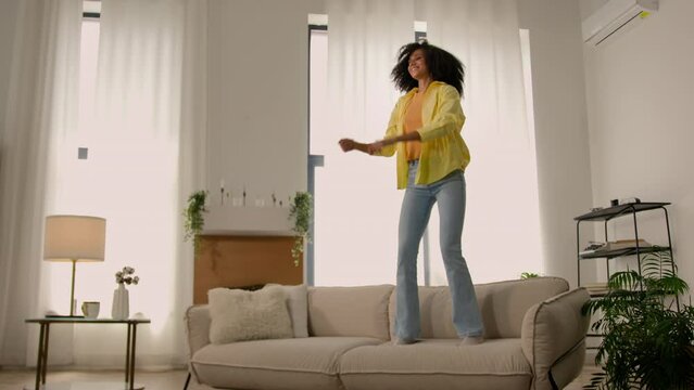 Active funny crazy African American girl young happy woman jumping on sofa couch in living room excited female jump dance loud music home party rejoicing carefree joyful lady weekend vacation freedom