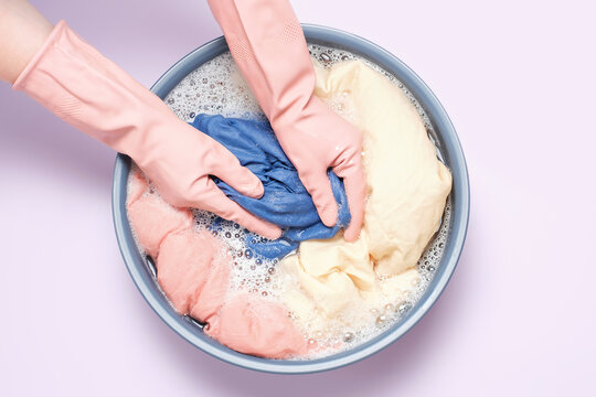 Female hands in rubber gloves washing clothes in basin on lilac background, top view.