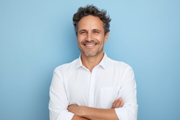 Portrait of handsome middle-aged man in white shirt, looking at camera and smiling, standing against blue background - obrazy, fototapety, plakaty