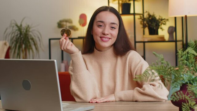 Caucasian young woman freelancer successful developer programmer holding two gold coins. Girl stock trader earning bitcoins after online monitoring trading operations sitting at home office at table