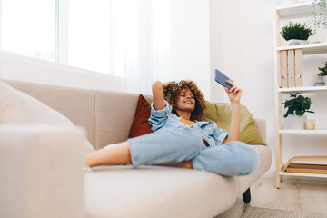 Cheerful woman sitting on a cozy couch at home, holding a mobile phone and smiling while chatting online.