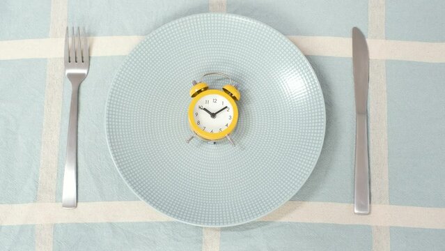 Empty plate with alarm clock and female hand on blue tablecloth background, intermittent fasting concept.