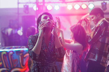 Young woman talking on smartphone and covering ear while partying in nightclub with loud music....