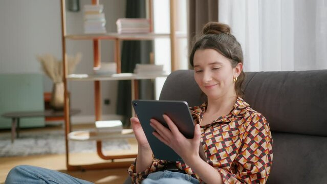 Smiling young caucasian woman sits in living room and uses tablet