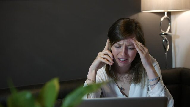 Sad and upset young professional woman receiving bad news on the phone at home working on laptop