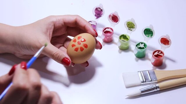Woman draws flower, leaves on beige boiled chicken egg. Female hand holds brush. Preparation for holiday of Happy Easter. Paints and paper on white background. Artist with red manicure. Springtime