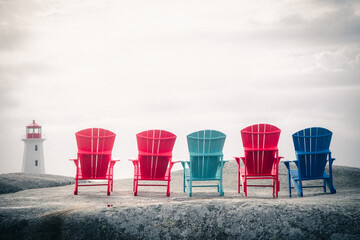 Row of red, blue and turquoise Adirondack chairs on the rocks in front of Peggy's Cove lighthouse in Nova Scotia, Canada - obrazy, fototapety, plakaty