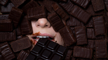The face of a caucasian woman surrounded by chocolates. The girl eats a bar of chocolate.