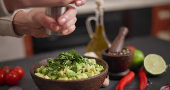 Salsa recipe - Woman pouring grinded pepper onto ingredients in wooden bowl - avocado, onion and cilantro