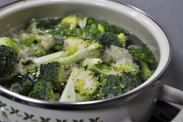 green soup with broccoli, onion, cauliflower, potatoes, peas boiling in a white pan on the stove in the kitchen
