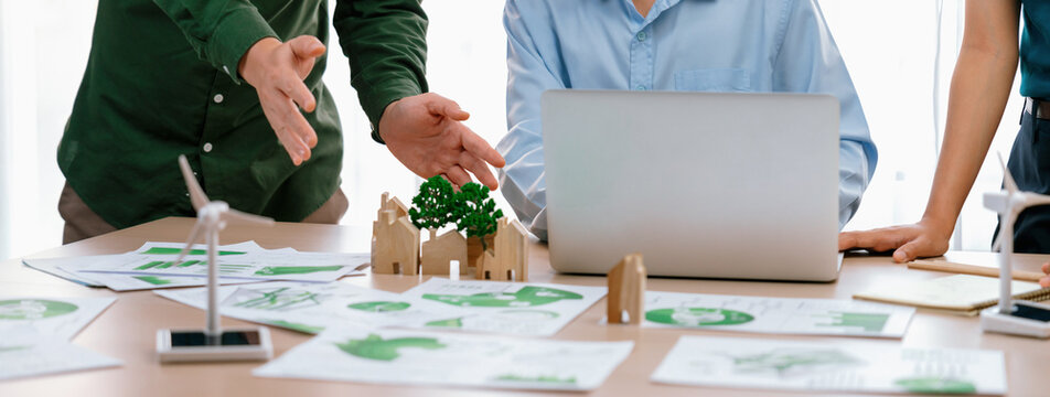 Cropped image of skilled businessman presented eco-friendly house project to general manager at meeting table with wooden block, windmill model and environmental document scatter around. Delineation.