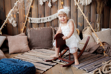 A little girl in a white lace dress is riding on a wooden horse swing in a wooden rustic interior
