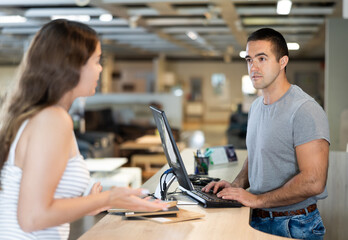 Focused young salesman of furniture showroom working with computer at information desk, advising...