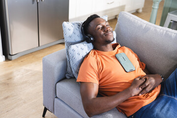 Smiling african american man on sofa in headphones, listening to music on smartphone, copy space