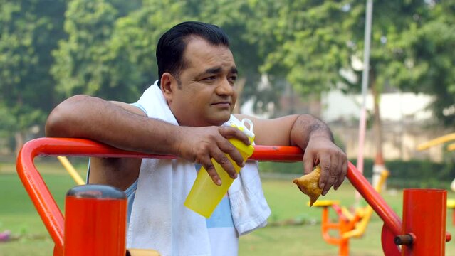 A tired Indian man with a samosa in hand - unhealthy food  workout  fitness  exercise  fat or fit  unsaturated fat food  confused. Obese adult having unhealthy fried snack after a tiring workout se...