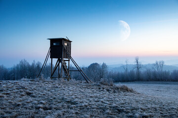 Naklejka premium Wooden hunting tower with frozen field on foreground and moon on night sky on background, winter landscape