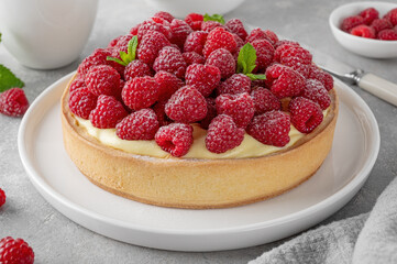 Delicious raspberry tart with vanilla custard cream sprinkled with powdered sugar on a white plate on a gray concrete background. Summer dessert.