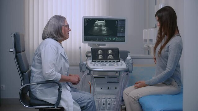 Caucasian woman sits it hospital sonography room, complains of abdominal pain to doctor. Medical specialist talks with female patient before examination. Ultrasound machine with monitor in background.
