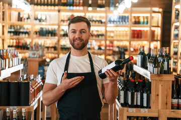 Man is holding bottle and smiling. Wine shop owner in white shirt and black apron