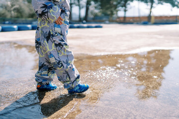 Small child in rubber boots and overalls walks through a puddle on the playground. Cropped. Faceless