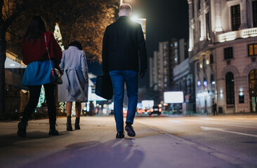 Back view of three individuals strolling together on a city sidewalk at night, with glowing festive...