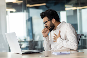 A bearded white-collar worker with eyewear coughing at desk with laptop.