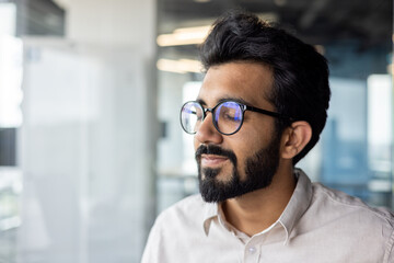 Man with black hair, glasses & beard smiles while looking out window - obrazy, fototapety, plakaty
