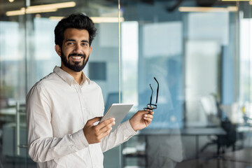 A bearded white-collar worker in glasses, holding a tablet and making a gesture - obrazy, fototapety, plakaty
