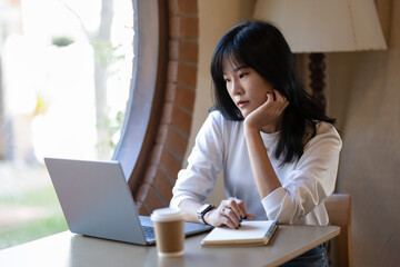 Pensive young adult woman working on a laptop in a cozy home setting, deep in thought..