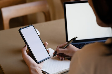 Businesswoman at a workspace with a smartphone and a laptop featuring a white blank screen, ideal for graphic mockups..