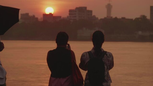 A safety volunteer standing and holding an umbrella watching two girls standing at the Hooghly River or Ganges riverbank clicking pictures of the beautiful sunset during monsoon or rainy season