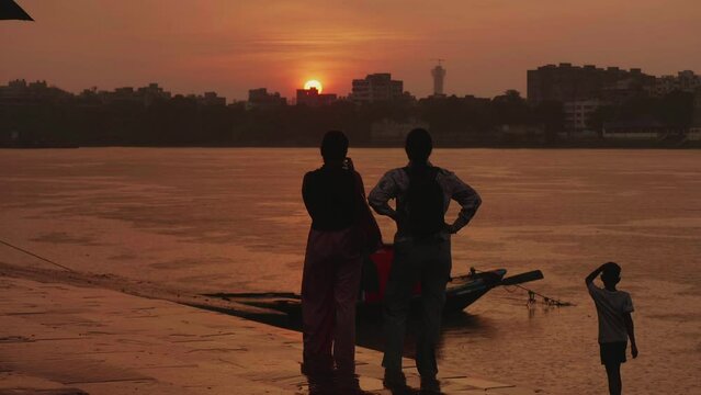 Two female travelers or tourists are standing alongside the Hooghly River or Ganges looking and clicking pictures or photos of the setting sun at the horizon or sunset in the evening in Kolkata.