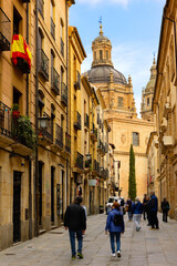 Picturesque cityscape of historic area of Salamanca overlooking people walking along typical narrow paved street leading to baroque building of La Clerecia with huge dome on sunny spring day, Spain.