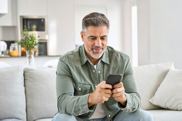 Older mature man using smartphone typing message relaxing on couch at home. Middle aged old male...