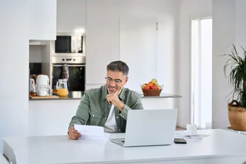 Fotobehang Happy middle aged mature man entrepreneur wearing eyeglasses looking at paper bill checking financial invoice or tax document making online payments working on laptop at home kitchen table. © insta_photos