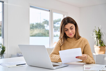 Middle aged older woman reading paper bill working at home using laptop computer in living room. Mature lady checking financial invoice or tax document making online payments working at home.