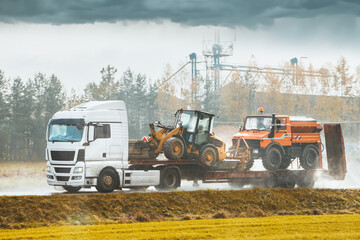 A white truck hauls heavy construction equipment on a trailer. Loaders and bulldozers are transported by a white truck ready for road construction.