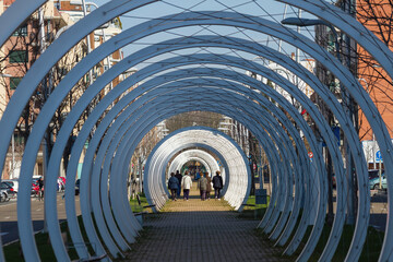 Adult women from behind walking in a group under white arches with metal braces that simulate a tunnel on a city avenue