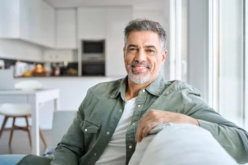 Tapeten Portrait of happy smiling middle aged mature senior 50 years old bearded man wearing green shirt sitting on couch at home interior looking at camera relaxing on sofa in modern house. © insta_photos