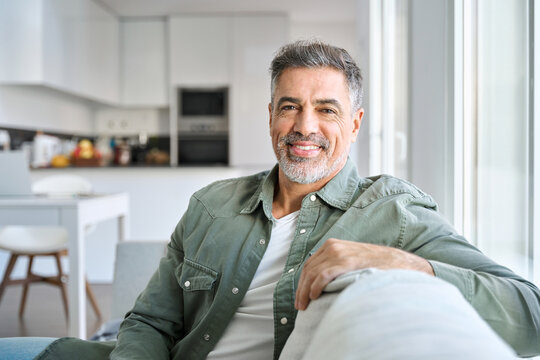Portrait of happy smiling middle aged mature senior 50 years old bearded man wearing green shirt sitting on couch at home interior looking at camera relaxing on sofa in modern house. - Powered by Adobe