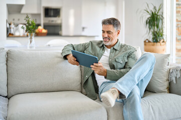 Happy middle aged man using digital tablet relaxing on couch at home. Happy mature male user...