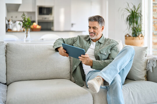 Happy middle aged man using digital tablet relaxing on couch at home. Happy mature male user holding tab computer browsing internet on pad technology device sitting on sofa in living room.