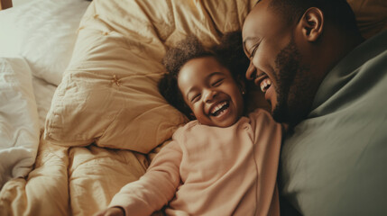 A joyful African American father and his young daughter share a heartfelt laugh while lying together in a cozy, sunlit bed - obrazy, fototapety, plakaty