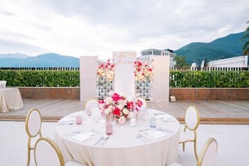Round festive table with chairs stands opposite a stand with bouquets of flowers. Caption: Love forever
