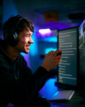 Low Key Lighting Shot Of Male Gamer In Front Of Computer Screens With Headphones And Controller