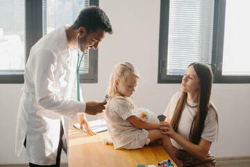 Fototapeta premium A preschool-aged girl with her mother is being examined by a pediatrician. A young man, a pulmonologist, conducts an examination using a phalendoscope.