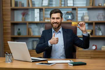 Smiling mature businessman in a sharp suit presenting a credit card while working from a neat home office setting.